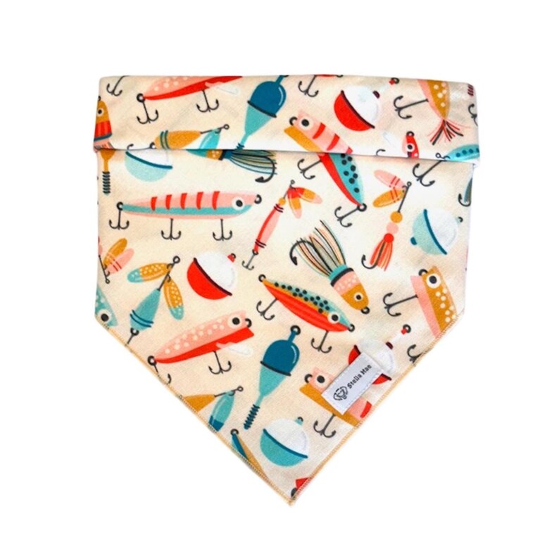 Puede incluir: Un pa&ntilde;uelo triangular para mascotas con fondo crema que presenta un patr&oacute;n colorido de se&ntilde;uelos de pesca, anzuelos y boyas en tonos rojos, azules, naranjas y amarillos. Una peque&ntilde;a etiqueta de tela blanca con el texto Stella Mae est&aacute; cosida.