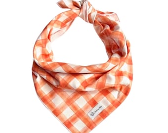 May include: A triangular pet bandana featuring an orange and white gingham check pattern. The fabric is tied in a knot at the top and includes a small white rectangular fabric label in the bottom corner with the text Stella Mae.