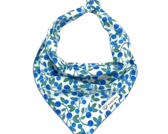 May include: A triangular pet bandana featuring a white background with a repeating pattern of blue blueberries and green leaves on stems. The fabric is tied at the top and includes a small white rectangular label with the text Stella Mae.