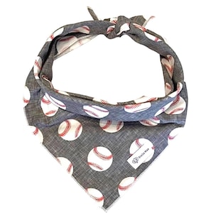 May include: A gray triangular pet bandana featuring a repeating pattern of white baseballs with red stitching. The fabric has a textured appearance and is tied at the top. A small white rectangular tag with the text Stella Mae is attached.