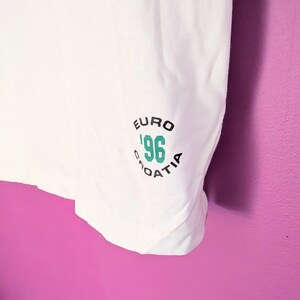 Puede incluir: Camiseta blanca con un gr&aacute;fico verde y negro que dice "EURO '96 CROATIA". El gr&aacute;fico es circular y presenta el a&ntilde;o '96 en color turquesa. La camiseta est&aacute; hecha de una tela blanca y suave.