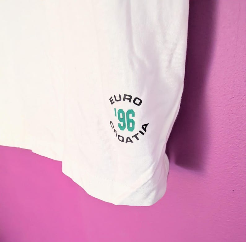 Puede incluir: Camiseta blanca con un gr&aacute;fico verde y negro que dice "EURO '96 CROATIA". El gr&aacute;fico es circular y presenta el a&ntilde;o '96 en color turquesa. La camiseta est&aacute; hecha de una tela blanca y suave.