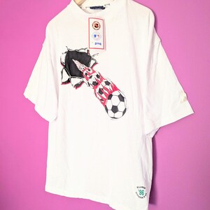 Puede incluir: Camiseta blanca con un dise&ntilde;o de bal&oacute;n de f&uacute;tbol saliendo de un agujero rasgado, con detalles rojos y negros. El texto "EURO '96 CROATIA" est&aacute; impreso. La prenda est&aacute; colgada en una percha de madera.