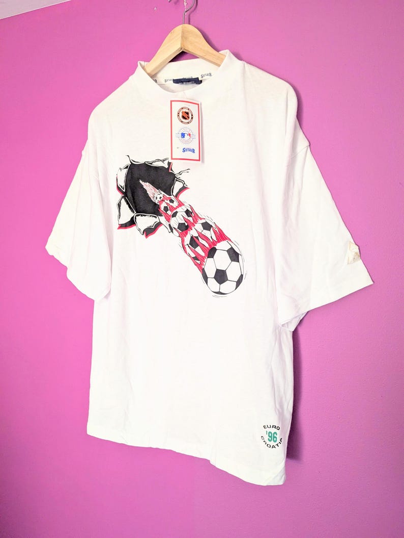 Puede incluir: Camiseta blanca con un dise&ntilde;o de bal&oacute;n de f&uacute;tbol saliendo de un agujero rasgado, con detalles rojos y negros. El texto "EURO '96 CROATIA" est&aacute; impreso. La prenda est&aacute; colgada en una percha de madera.