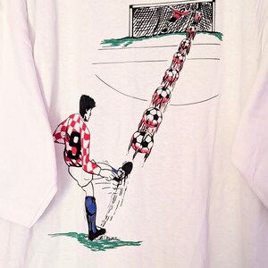 Puede incluir: Camiseta blanca con una ilustraci&oacute;n de dibujos animados de un jugador de f&uacute;tbol pateando un bal&oacute;n hacia una porter&iacute;a. El jugador lleva una camiseta a cuadros rojos y blancos con el n&uacute;mero 9. El bal&oacute;n de f&uacute;tbol se representa en movimiento, con un rastro de balones que conducen a la porter&iacute;a.
