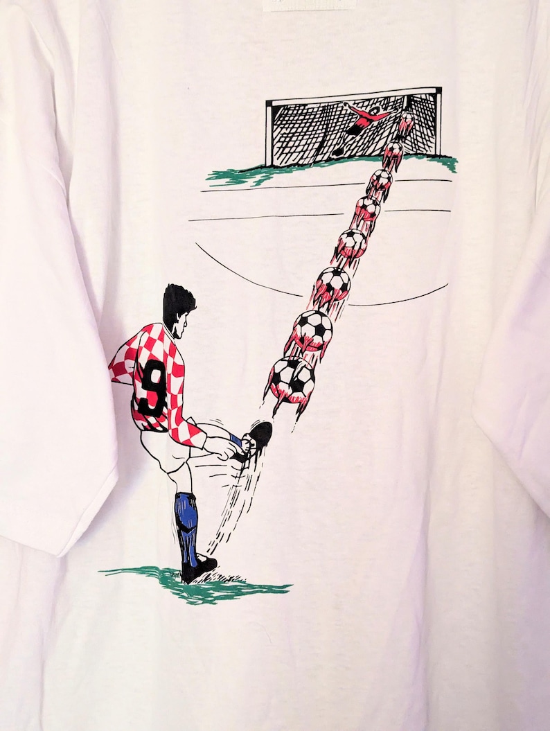 Puede incluir: Camiseta blanca con una ilustraci&oacute;n de dibujos animados de un jugador de f&uacute;tbol pateando un bal&oacute;n hacia una porter&iacute;a. El jugador lleva una camiseta a cuadros rojos y blancos con el n&uacute;mero 9. El bal&oacute;n de f&uacute;tbol se representa en movimiento, con un rastro de balones que conducen a la porter&iacute;a.