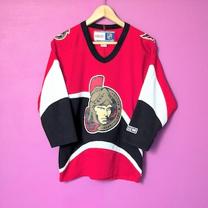 May include: A red, black, and white hockey jersey with the Ottawa Senators logo. The logo features a gold helmet with a warrior's face and a gold laurel wreath. The jersey has a V-neck and long sleeves.