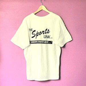 May include: White t-shirt with a black print that reads "The Sports Link Inc. "Soccer Starts Here..."