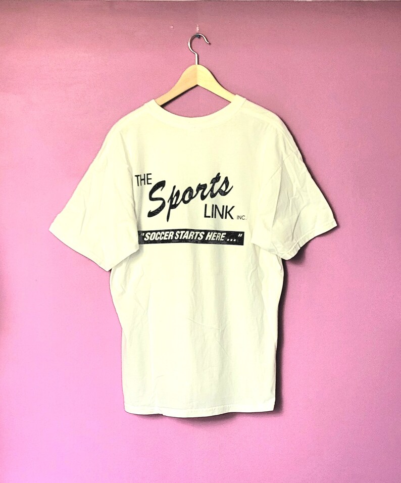May include: White t-shirt with a black print that reads "The Sports Link Inc. "Soccer Starts Here..."