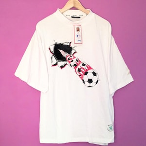 Puede incluir: Camiseta blanca con un dise&ntilde;o de bal&oacute;n de f&uacute;tbol rompiendo un agujero rasgado, con detalles en rojo y negro. La camiseta tiene cuello redondo, mangas cortas y una etiqueta. El texto 'EURO '96 CROATIA' est&aacute; impreso en la parte inferior.
