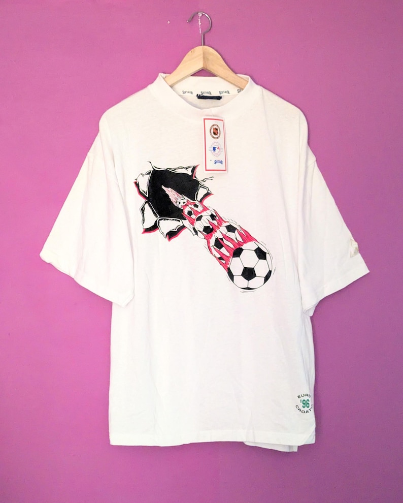 Puede incluir: Camiseta blanca con un dise&ntilde;o de bal&oacute;n de f&uacute;tbol rompiendo un agujero rasgado, con detalles en rojo y negro. La camiseta tiene cuello redondo, mangas cortas y una etiqueta. El texto 'EURO '96 CROATIA' est&aacute; impreso en la parte inferior.