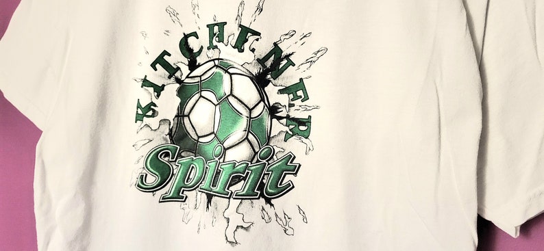 May include: White t-shirt featuring a green and white football graphic with the text "Kitchener Spirit".