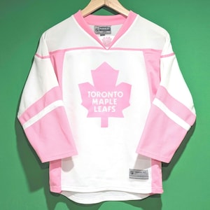 May include: A white and pink Reebok hockey jersey featuring the Toronto Maple Leafs logo in pink on the chest. The jersey has a V-neck collar, long sleeves with pink and white color-blocking, and a Reebok brand tag on the lower front hem.