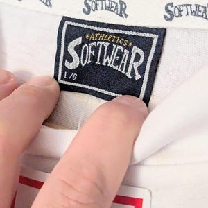 Puede incluir: Primer plano de una etiqueta de camiseta blanca con la palabra "SOFTWEAR" impresa en una fuente estilizada. La etiqueta es azul marino con texto blanco y las palabras "ATHLETICS" y "L/G". El cuello de la camisa tambi&eacute;n tiene la palabra "SOFTWEAR" impresa.