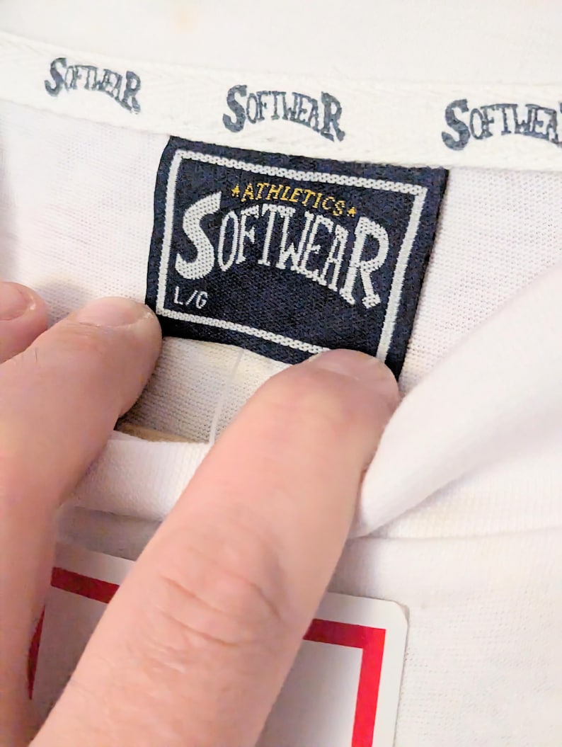 Puede incluir: Primer plano de una etiqueta de camiseta blanca con la palabra "SOFTWEAR" impresa en una fuente estilizada. La etiqueta es azul marino con texto blanco y las palabras "ATHLETICS" y "L/G". El cuello de la camisa tambi&eacute;n tiene la palabra "SOFTWEAR" impresa.