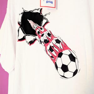 Puede incluir: Camiseta blanca con un gr&aacute;fico de bal&oacute;n de f&uacute;tbol que atraviesa un agujero rasgado. El dise&ntilde;o incluye balones de f&uacute;tbol en blanco y negro con llamas rojas y una etiqueta con texto.
