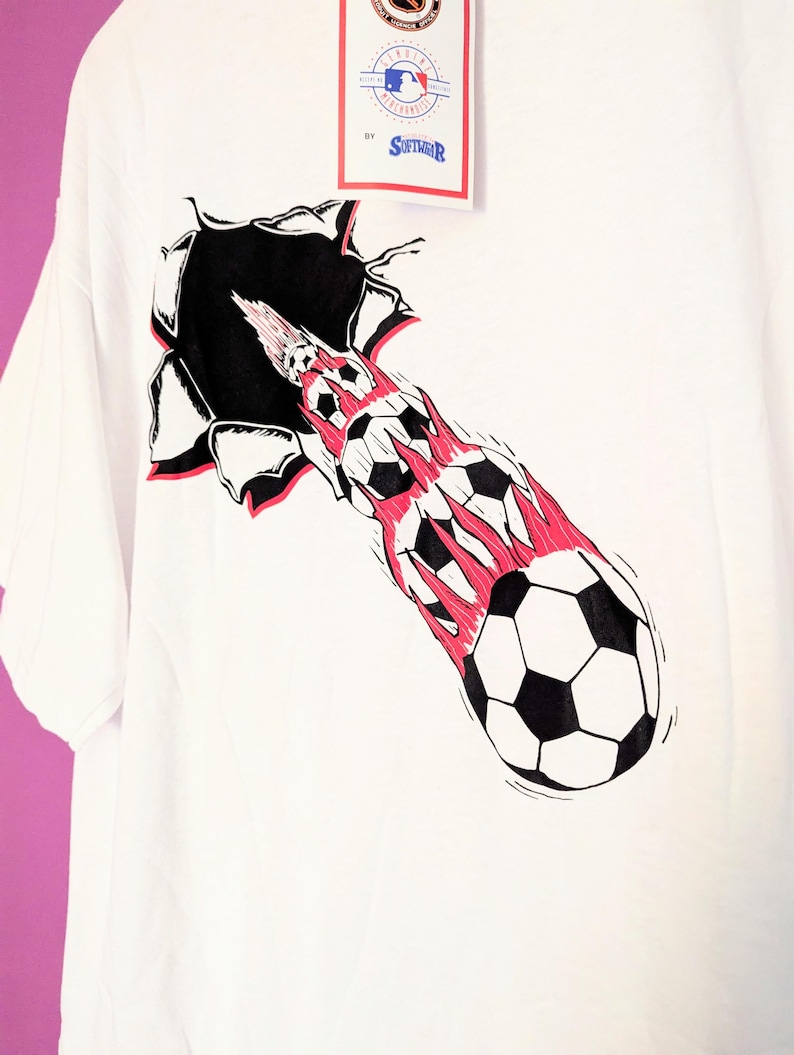 Puede incluir: Camiseta blanca con un gr&aacute;fico de bal&oacute;n de f&uacute;tbol que atraviesa un agujero rasgado. El dise&ntilde;o incluye balones de f&uacute;tbol en blanco y negro con llamas rojas y una etiqueta con texto.