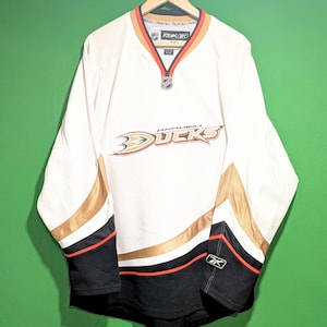 May include: A white Anaheim Ducks hockey jersey featuring a V-neck collar with orange and black trim. The front displays the team logo and text, with gold, black, and orange stripes on the sleeves and hem. A Reebok logo is on the left sleeve cuff.
