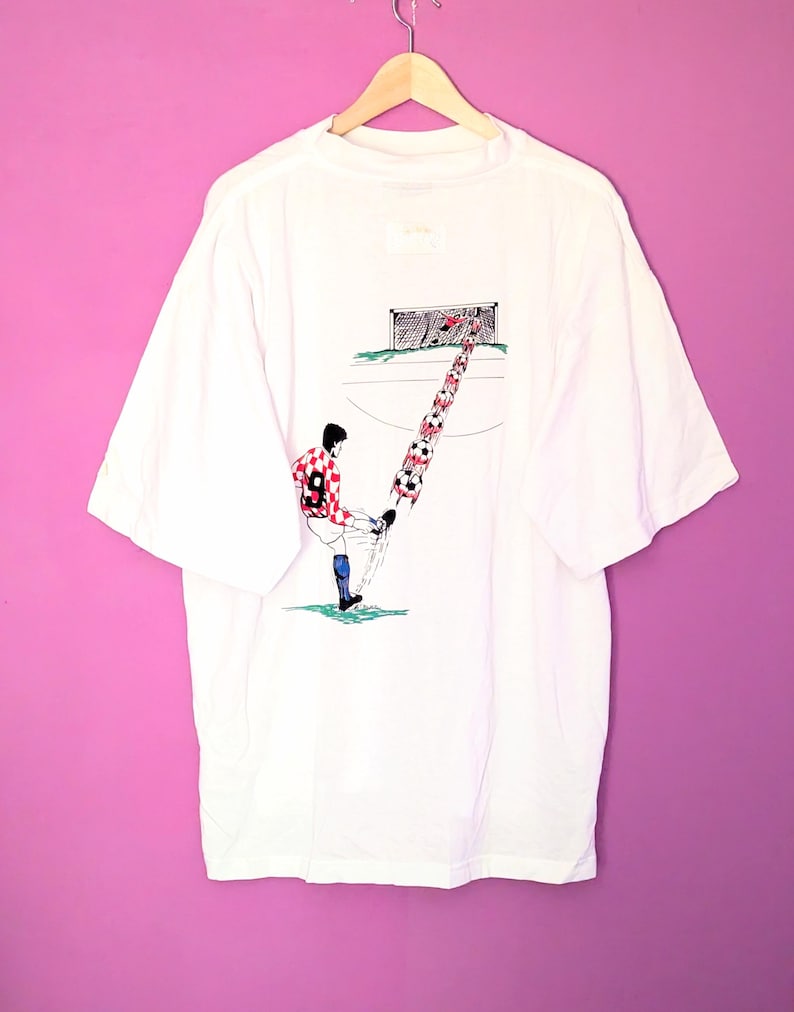 Puede incluir: Camiseta blanca con un dise&ntilde;o de f&uacute;tbol. La ilustraci&oacute;n muestra a un jugador pateando un bal&oacute;n, con una serie de balones apilados hacia la porter&iacute;a. El jugador lleva un uniforme a cuadros rojos y blancos. La camiseta est&aacute; colgada en una percha de madera.