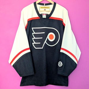 May include: A vintage black hockey jersey with white and red accents. The jersey features the Philadelphia Flyers logo on the front and is hanging on a wooden hanger. The sleeves are white with red stripes.