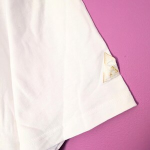Puede incluir: Camiseta blanca de manga corta con un peque&ntilde;o parche bordado en la manga. El parche es de color beige claro con costuras doradas. La camiseta est&aacute; hecha de una tela blanca suave. El fondo es de color morado liso.