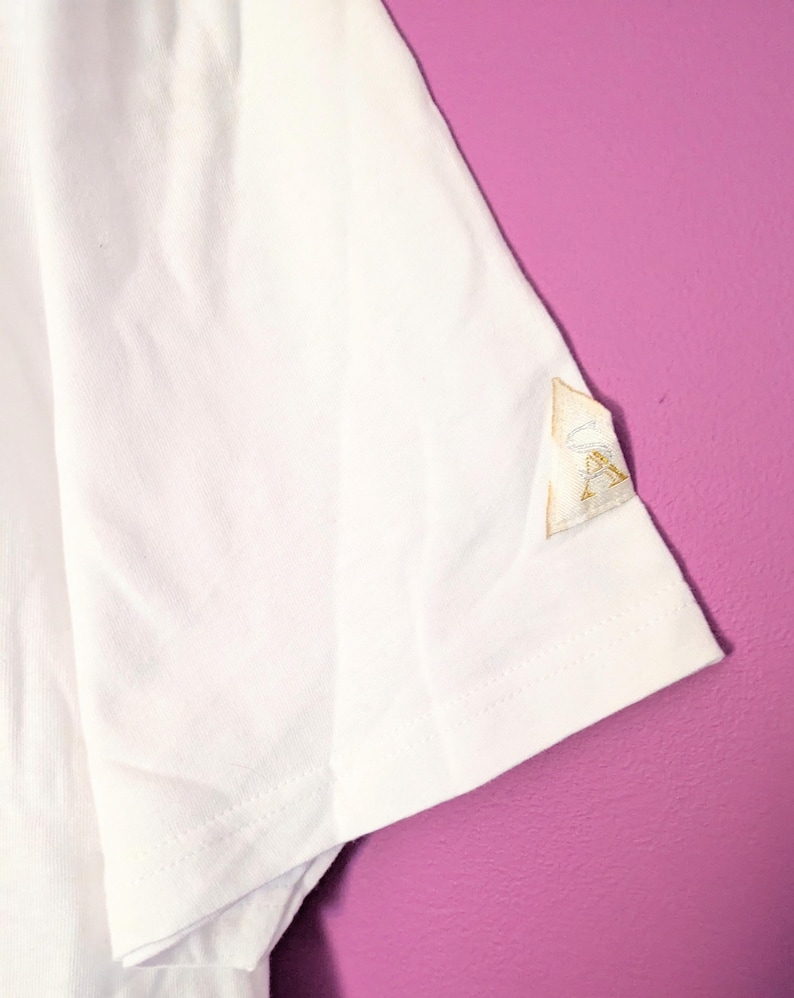 Puede incluir: Camiseta blanca de manga corta con un peque&ntilde;o parche bordado en la manga. El parche es de color beige claro con costuras doradas. La camiseta est&aacute; hecha de una tela blanca suave. El fondo es de color morado liso.