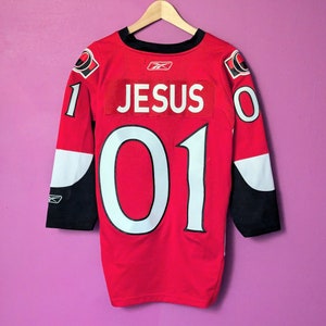 May include: A red hockey jersey with black and white accents. The number "01" is printed on the back in white with a black outline. The name "JESUS" is printed above the number in white. The jersey is hanging on a hanger.