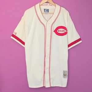 May include: White baseball jersey with red trim and the Cincinnati Reds logo on the front. The logo is a red 'C' with a red 'REDS' inside. The jersey has a button-up front and short sleeves.