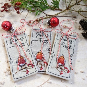 May include: Three embroidered Christmas gift tags with a robin design and the words "Frohes Fest". Each tag is made of a light-colored fabric, adorned with red and white twine, and topped with a red polka-dot ornament.