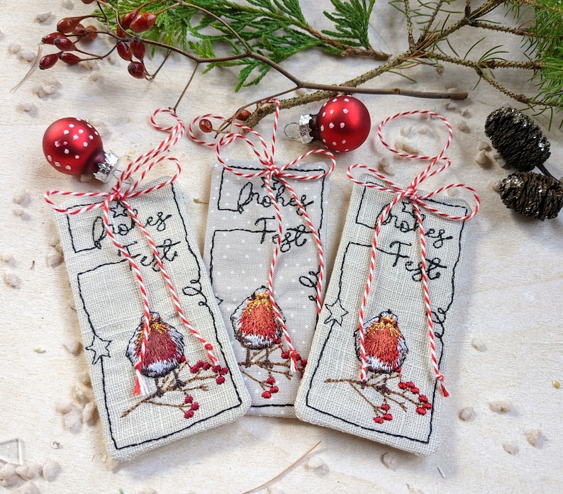May include: Three embroidered Christmas gift tags with a robin design and the words "Frohes Fest". Each tag is made of a light-colored fabric, adorned with red and white twine, and topped with a red polka-dot ornament.