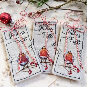 May include: Three rectangular Christmas ornaments with embroidered robin bird designs, the text "Frohes Fest", and red and white twine bows. A red and white polka-dot ornament and sprigs of berries are in the background.