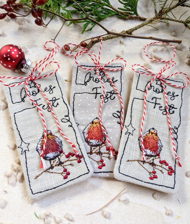 May include: Three rectangular Christmas ornaments with embroidered robin bird designs, the text "Frohes Fest", and red and white twine bows. A red and white polka-dot ornament and sprigs of berries are in the background.