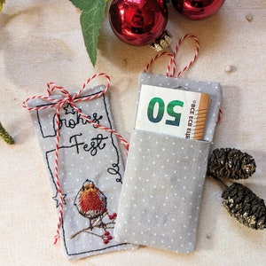 May include: Two gray fabric gift card holders with white polka dots. One has a red and white string tie and embroidered text and a bird. The other holds a 50 Euro bill. Christmas ornaments are in the background.