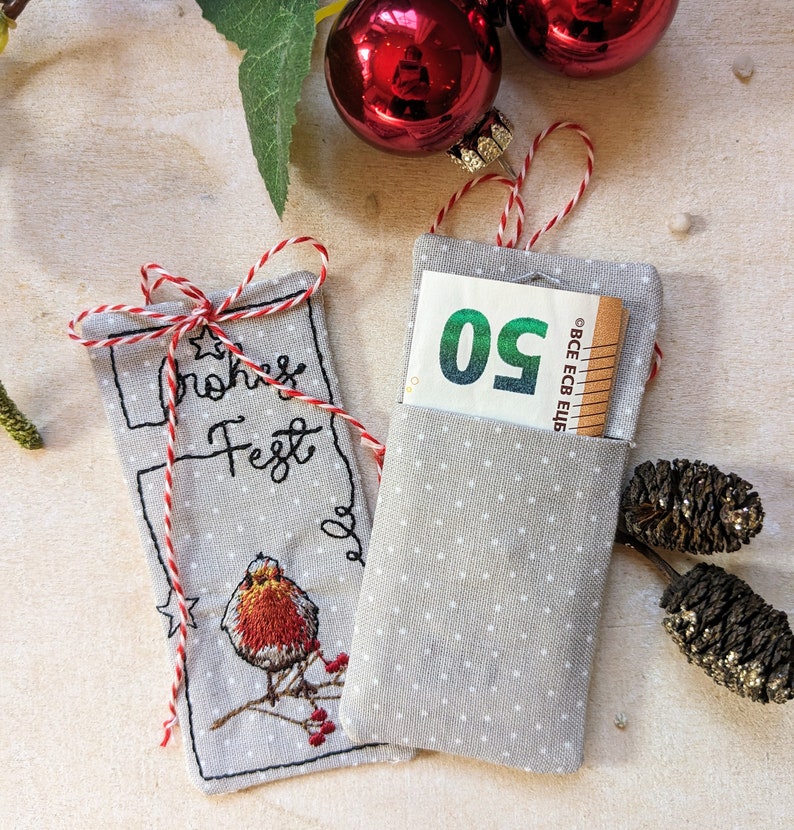 May include: Two gray fabric gift card holders with white polka dots. One has a red and white string tie and embroidered text and a bird. The other holds a 50 Euro bill. Christmas ornaments are in the background.