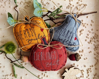 May include: Three stuffed fabric ornaments shaped like pumpkins and a pear. The orange pumpkin features the text 'HERBST zauber', the red pumpkin says 'LIEBLINGS zeit', and the grey pear is embroidered with 'HERBST geflüster'. Each has twine.