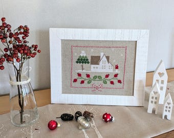 May include: A white framed cross-stitch of a winter scene with a small house, a tree, and red berries. The cross-stitch is on a white background with a red border. The frame is on a table with a white tablecloth and a vase of red berries.