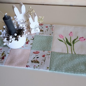 Könnte beinhalten: Ein weißer Tischläufer mit Patchwork-Muster aus grünen und rosa Quadraten. Der Läufer hat ein weißes Mittelquadrat mit einem gestickten Muster aus rosa Tulpen. Der Läufer ist mit weißen Holzhasenfiguren und einer schwarzen Kerze verziert.