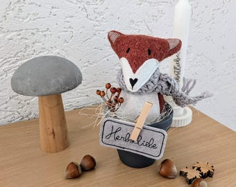 May include: A plush fox with a rust-colored head and white face, wearing a gray knit scarf, sits in a dark gray pot. A light gray fabric tag attached with a wooden clip reads "Herbstliebe". Nearby are a concrete mushroom, hazelnuts, and a white candle.