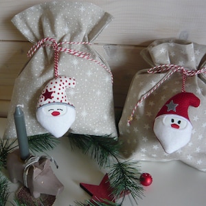 Könnte beinhalten: Zwei beigefarbene Geschenktüten aus Stoff mit weißen Sternenmustern, jeweils mit einer rot-weiß gestreiften Kordel gebunden. An jeder Tasche befindet sich ein Plüsch-Weihnachtsmann-Anhänger mit weißem Bart und einer roten Mütze.