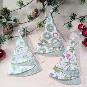 Könnte beinhalten: Drei handgefertigte Weihnachtsbaumornamente mit mehrschichtigen Stoffdesigns. Jeder Baum hat ein anderes Muster: botanisch, Kranz und floral. Die Bäume sind mit Bindfäden und weißen Perlen zum Aufhängen verziert. Rote Kugeln und Tannenzweige vervollständigen die festliche Szene.