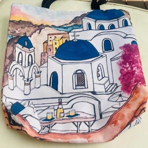 May include: A white canvas tote bag with a colorful illustration of a Greek village. The illustration features white buildings with blue accents, a pink tree, and a table with wine glasses and a bottle of wine.