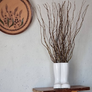 Può includere: Un vaso bianco a forma di stivale in ceramica pieno di rami marroni e sottili. Un piatto decorativo rotondo e marrone con disegni di grano e fiori è appeso al muro. Il vaso è appoggiato su una mensola di legno contro un muro bianco strutturato.