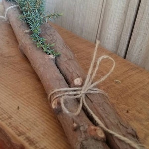 Puede incluir: Dos troncos de madera rústicos atados con cuerda, adornados con ramitas de follaje azul-verdoso. Una pequeña etiqueta con las palabras "Thank You!" está adjunta. El arreglo descansa sobre una superficie de madera.