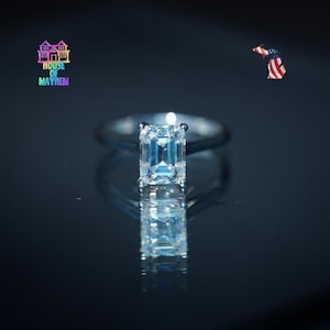 May include: A silver-toned ring featuring a clear, rectangular emerald-cut gemstone set in a four-prong setting. The ring is positioned against a dark background, creating a clear reflection below it. The text House of Mayhem is visible.