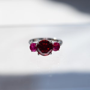 May include: A silver ring featuring a large, faceted, deep red gemstone flanked by two smaller, round, magenta gemstones. The ring is set against a bright white background, highlighting the vibrant colors of the stones.