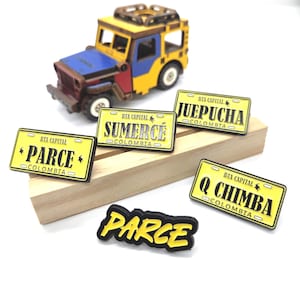 May include: A wooden toy jeep in yellow, blue, and red, with a set of yellow license plate-shaped magnets. The magnets have black text that reads "PARCE", "SUMERCE", "JUEPUCHA", and "Q CHIMBA", with "BTA CAPITAL COLOMBIA" on each.