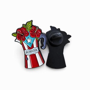Pode incluir: Um alfinete de esmalte colorido em forma de cafeteira, com a bandeira de Porto Rico e flores de hibisco vermelhas. O alfinete tem um verso preto e a palavra "BORICUA" est&aacute; impressa na frente.