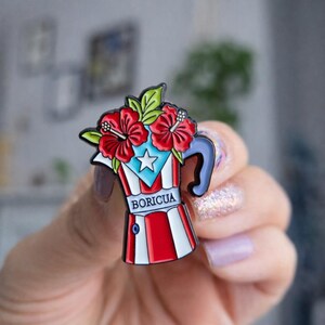 Pode incluir: Um alfinete de esmalte colorido em forma de cafeteira, com o design da bandeira de Porto Rico. O alfinete tem listras vermelhas e brancas, uma estrela azul e a palavra "BORICUA". &Eacute; coberto com flores de hibisco vermelhas e folhas verdes.