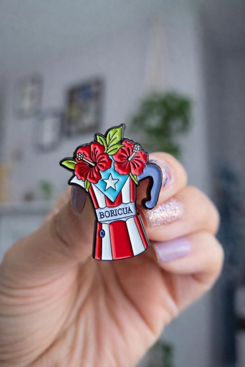 Pode incluir: Um alfinete de esmalte colorido em forma de cafeteira, com o design da bandeira de Porto Rico. O alfinete tem listras vermelhas e brancas, uma estrela azul e a palavra "BORICUA". &Eacute; coberto com flores de hibisco vermelhas e folhas verdes.