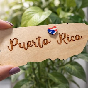 May include: A wooden cutout in the shape of Puerto Rico, with the words "Puerto Rico" engraved in a cursive font. A heart-shaped pin with the Puerto Rican flag is attached. The cutout is held in front of a leafy green background.